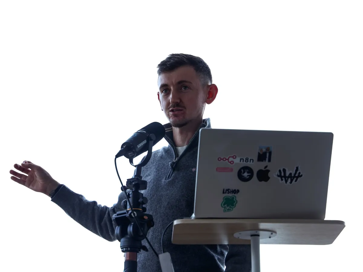 Roman Rodomansky speaking into a microphone and gesturing with one hand, his laptop with n8n stickers on the lectern.