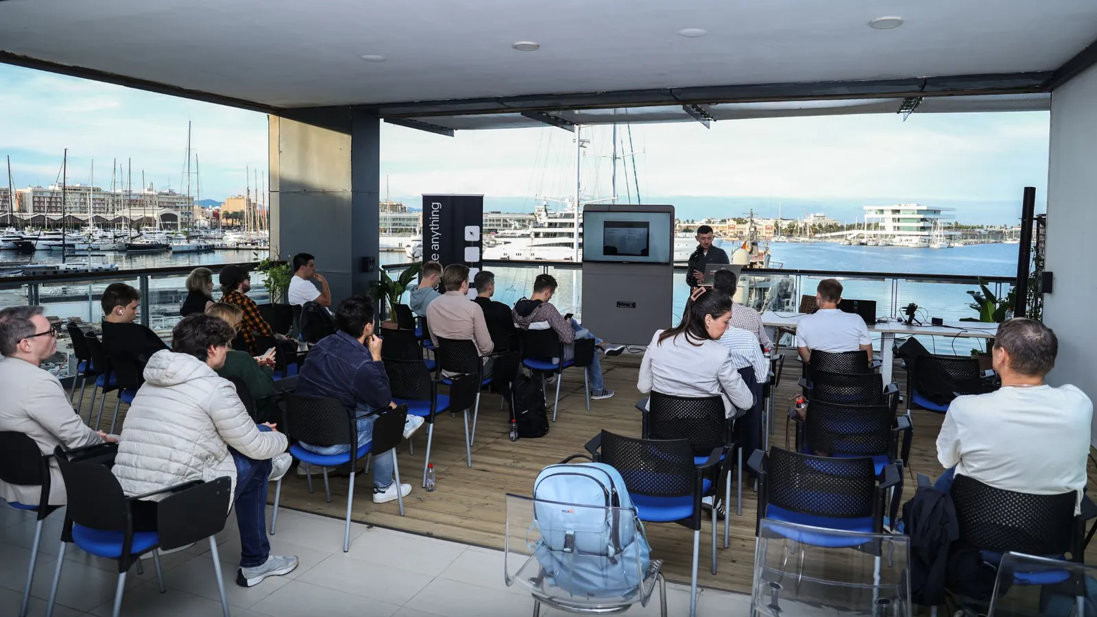 The n8n Valencia workshop on Base 2's covered terrace: people on blue chairs facing the speaker and a screen, with yachts and the port of Valencia behind.