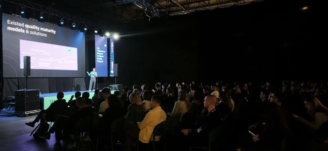The audience in a dark hall facing the stage and a slide on existing quality maturity models.